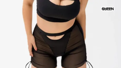Woman wearing a black crop top and sheer black shorts with cutouts, standing against a white background; QUEEN logo in the top-right corner.