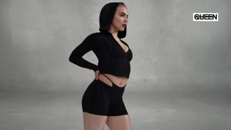 Pole Dance Shorts How to Choose Coverage Cut and Grip Model in a black zip-up cropped hoodie and matching shorts, hands on hips, gray studio backdrop, QUEEN logo in corner.