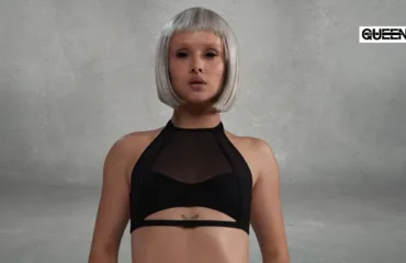 Pole Dance Tops How to Choose Support Fit and Style Woman with a short platinum bob and a black mesh-ctop, facing the camera, butterfly tattoo under the bust, gray textured backdrop with a Queen logo in the corner.