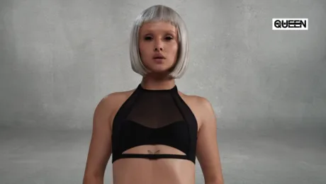 Pole Dance Tops How to Choose Support Fit and Style Woman with a short platinum bob and a black mesh-ctop, facing the camera, butterfly tattoo under the bust, gray textured backdrop with a Queen logo in the corner.