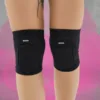 Two black knee pads worn on both knees, brand text 'QUEEN' visible, with pink paint splash background (product photo)