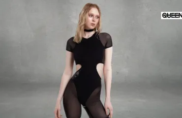 Pole Dance Bodysuit Guide When to Wear It and How to Choose Woman model wearing a black cut-out bodysuit with sheer panels poses against a gray studio wall; a QUEEN logo visible in the upper-right corner.