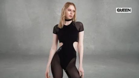 Pole Dance Bodysuit Guide When to Wear It and How to Choose Woman model wearing a black cut-out bodysuit with sheer panels poses against a gray studio wall; a QUEEN logo visible in the upper-right corner.