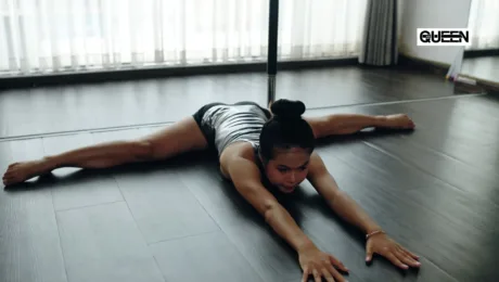 Pole Dance Wear for Beginners What to Buy First Woman in a gray outfit stretching into a full split on a dark wooden studio floor beside a vertical dance pole, with sheer curtains and a 'QUEEN' logo in the corner.