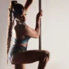 Pole dancer in teal sports bra and dark shorts, gripping a vertical pole with a long braided ponytail, in a neutral studio.QUEEN logo in the corner